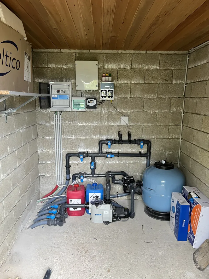 Local technique de piscine dans un abri en bois ouvert, avec filtre bleu, pompe et vannes