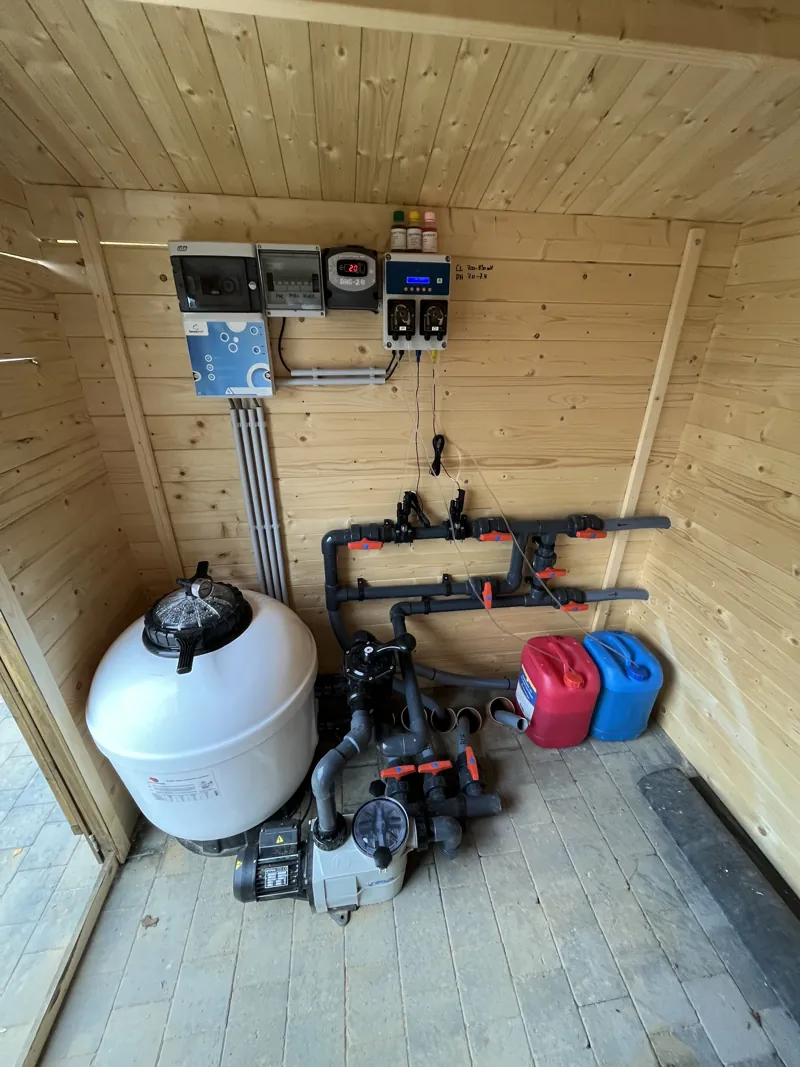 Local technique de piscine dans un chalet en bois, avec filtre blanc, pompe et vannes
